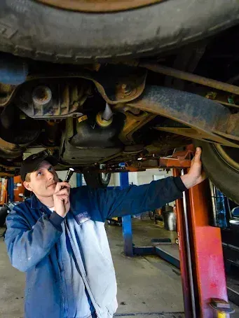 A technician in a blue and gray uniform inspects the undercarriage of a vehicle lifted on a hydraulic rack in a garage.