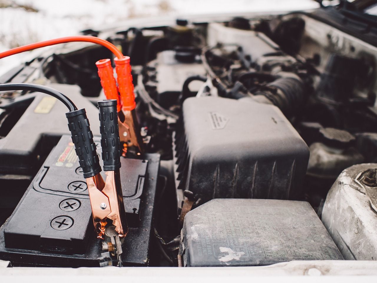 Car engine bay with black and red jumper cables attached to a battery terminal for a jump-start.