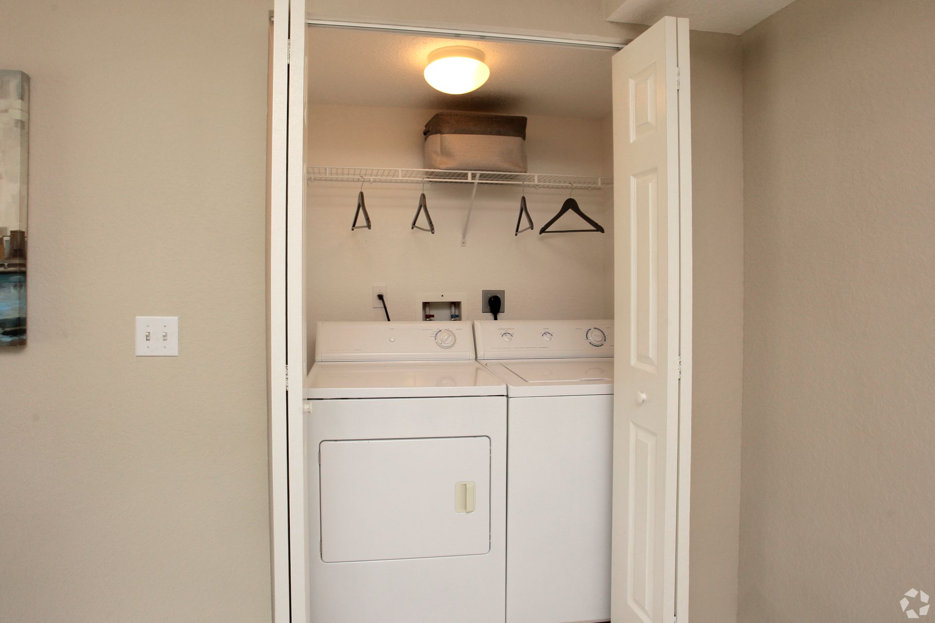 Laundry closet with stacked washer and dryer, white shelves, and overhead light in a beige hallway 4120 at Palm Beach Gardens Apartments.