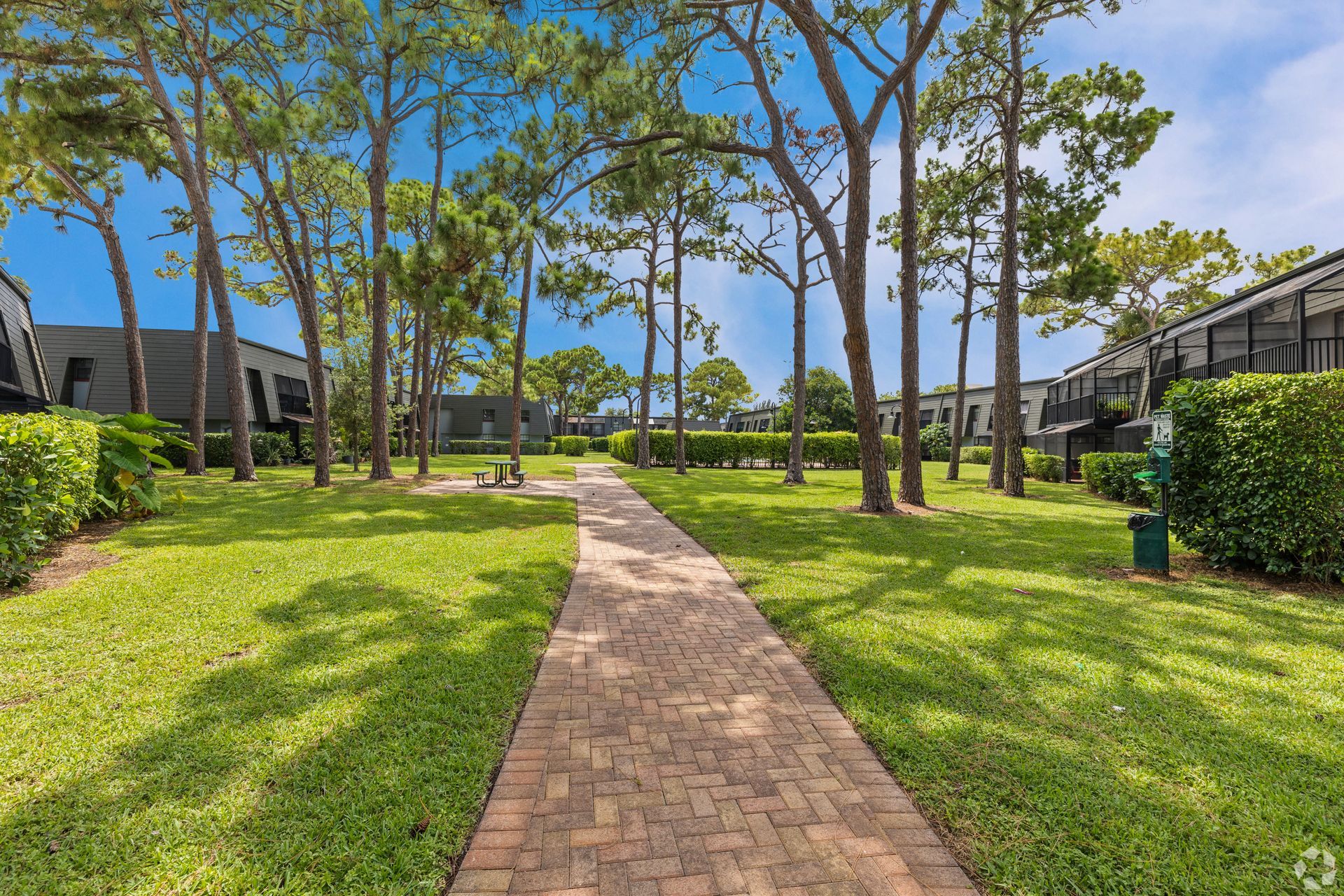 Brick walkway through a green courtyard with trees and apartment buildings under a blue sky at 4120 at Palm Beach Gardens Apartments in FL.