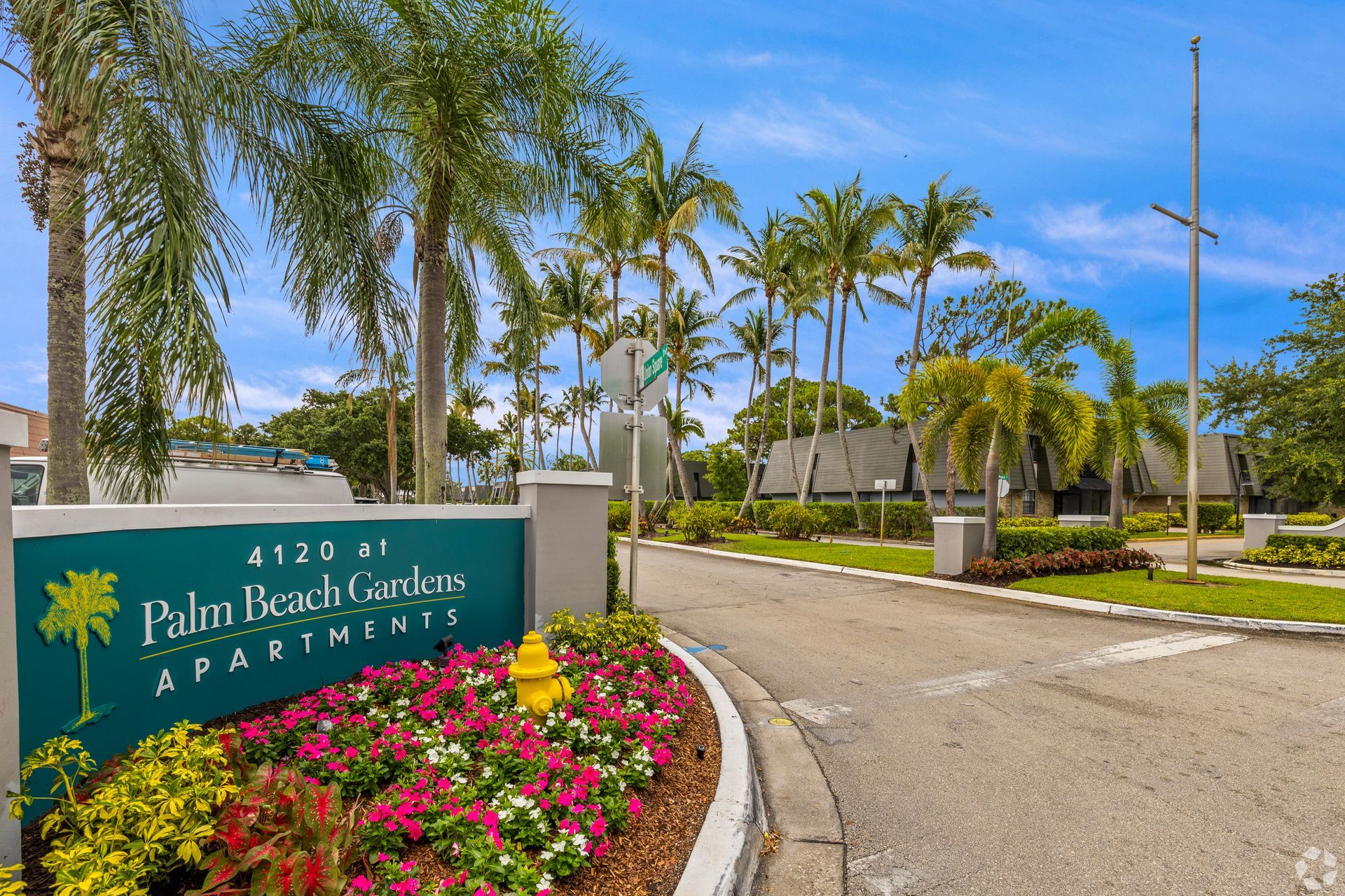 Entrance sign for Pula Bahia Gardens Apartments with palm trees, flowers, and a driveway under a blue sky at 4120 at Palm Beach Gardens Apartments in FL.