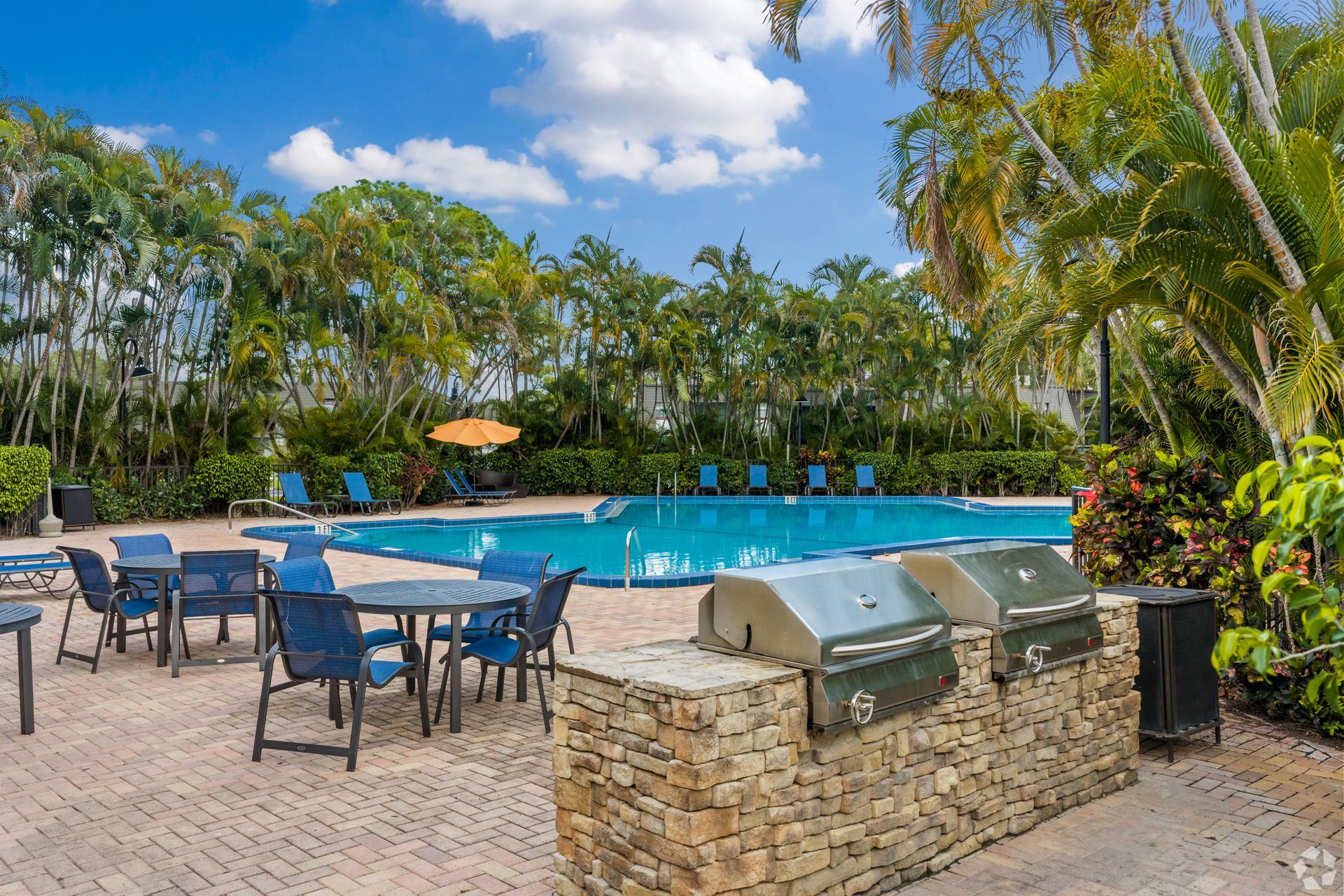 Outdoor pool with lounge chairs, patio tables, and a stone grill area surrounded by palm trees at 4120 at Palm Beach Gardens Apartments in FL