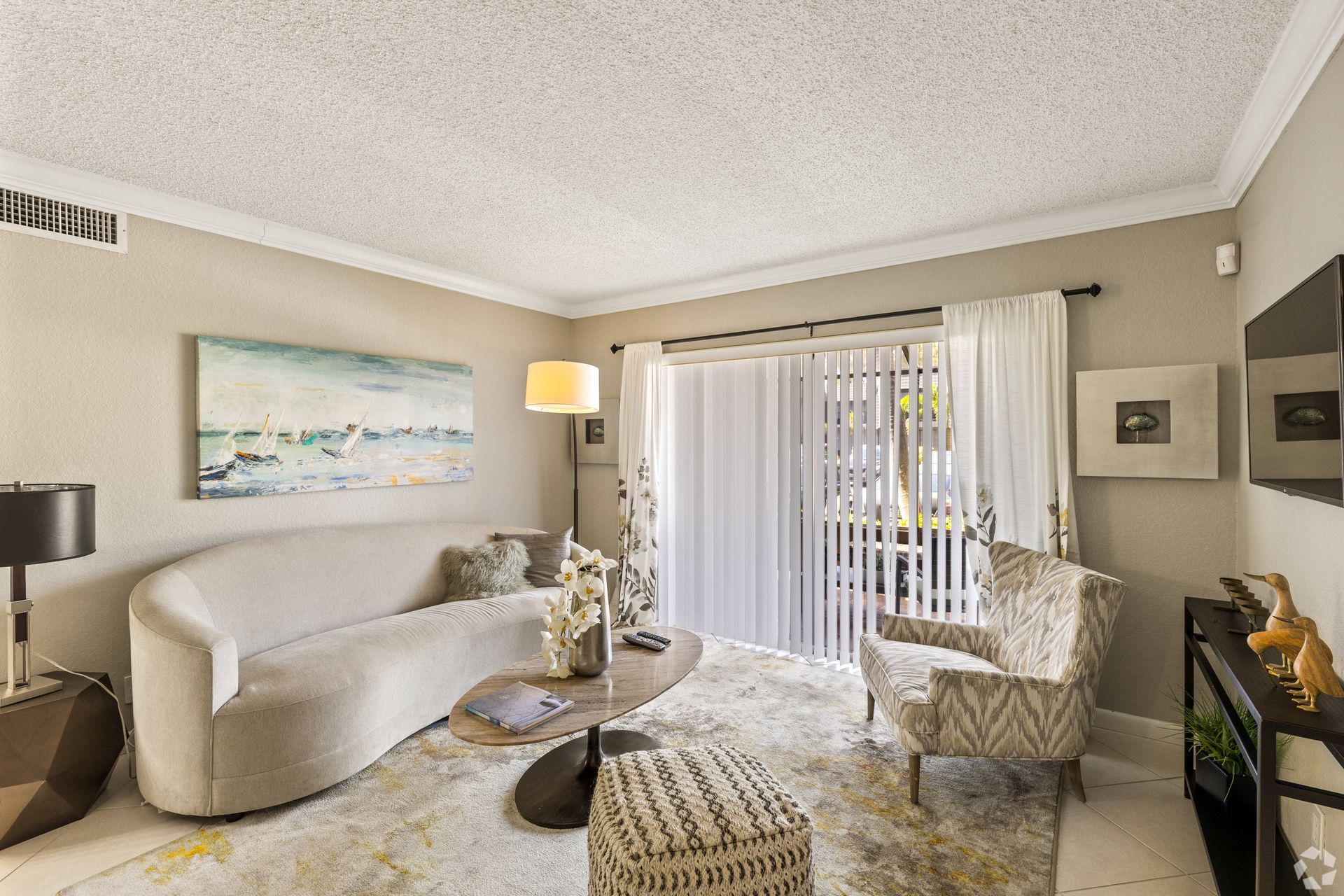 Bright living room with beige sofa, patterned chairs, floor lamp, artwork, and vertical blinds by a sliding door at 4120 at  Palm Beach Gardens Apartments in  FL.