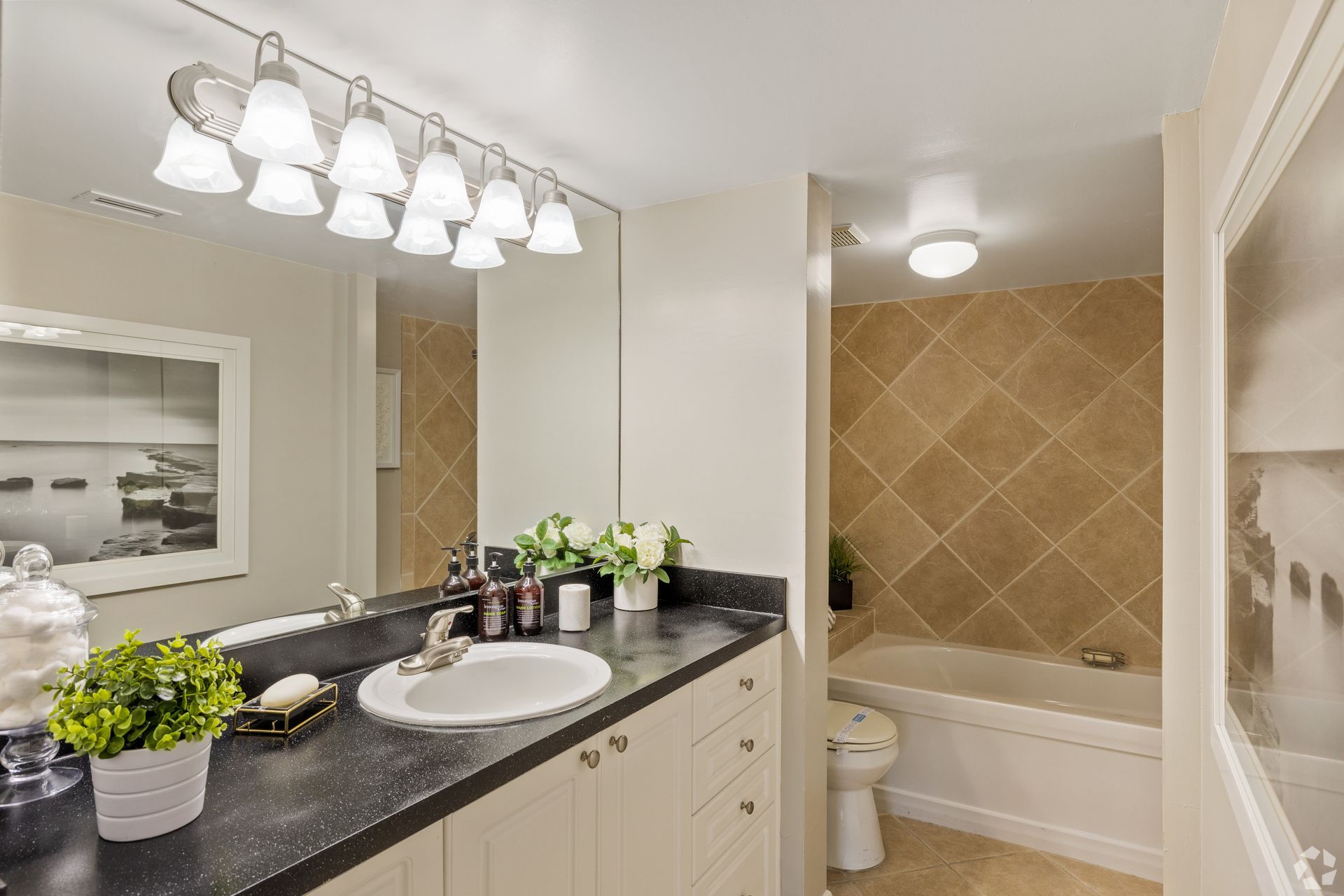 Bright bathroom vanity with black countertop, white cabinets, and a tub with beige tile surround at 4120 at Palm Beach Gardens Apartments in FL