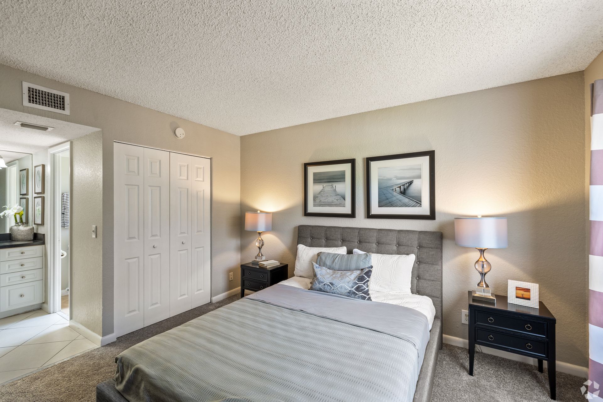 Bright bedroom with a gray bed, bedside lamps, framed art, and an open doorway to a bathroom at 4120 at  Palm Beach Gardens Apartments in  FL