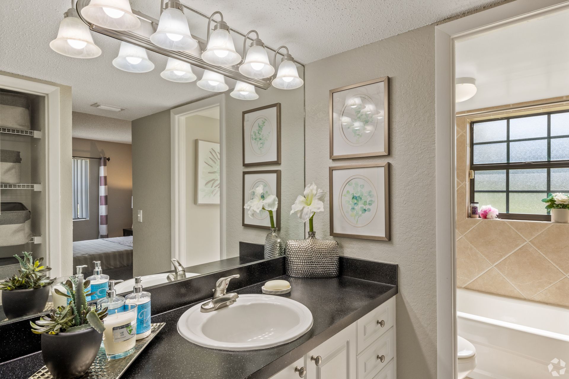Bright bathroom vanity with double sinks, black countertop, framed art, and a windowed shower area at 4120 at Palm Beach Gardens Apartments in FL