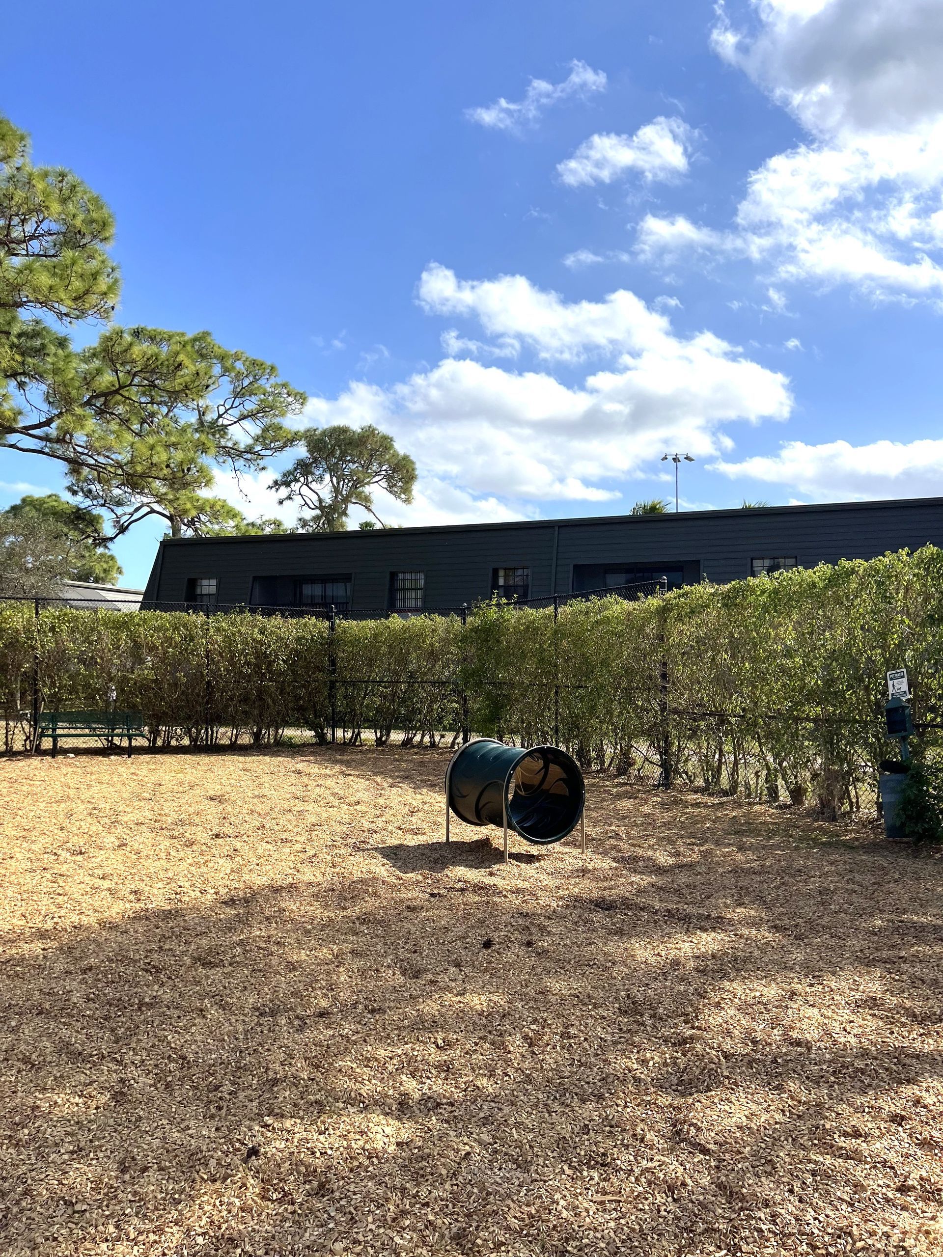 Black barrel on wood chips in a sunny outdoor yard with hedges, trees, and a long dark building at 4120 at Palm Beach Gardens Apartments