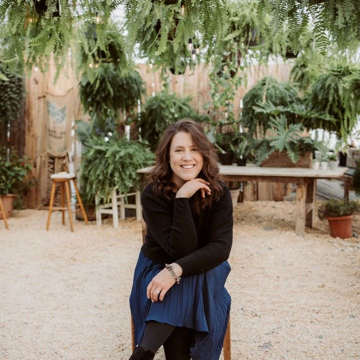 Woman smiling, sitting in a greenhouse, surrounded by plants and wooden furniture.