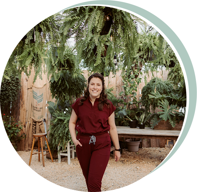Woman in burgundy scrubs stands in a greenhouse, surrounded by plants.