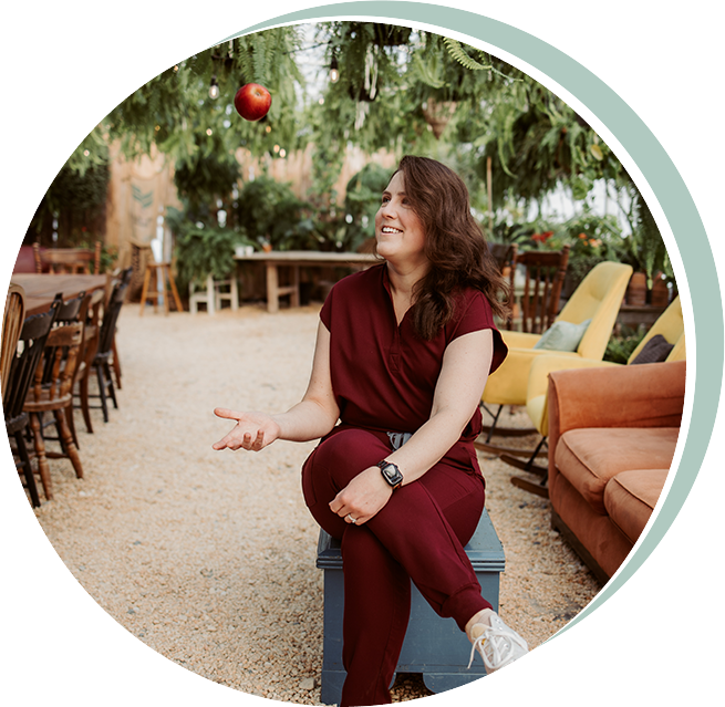Woman in red scrubs, tossing an apple, smiling in a garden with tables and seating.