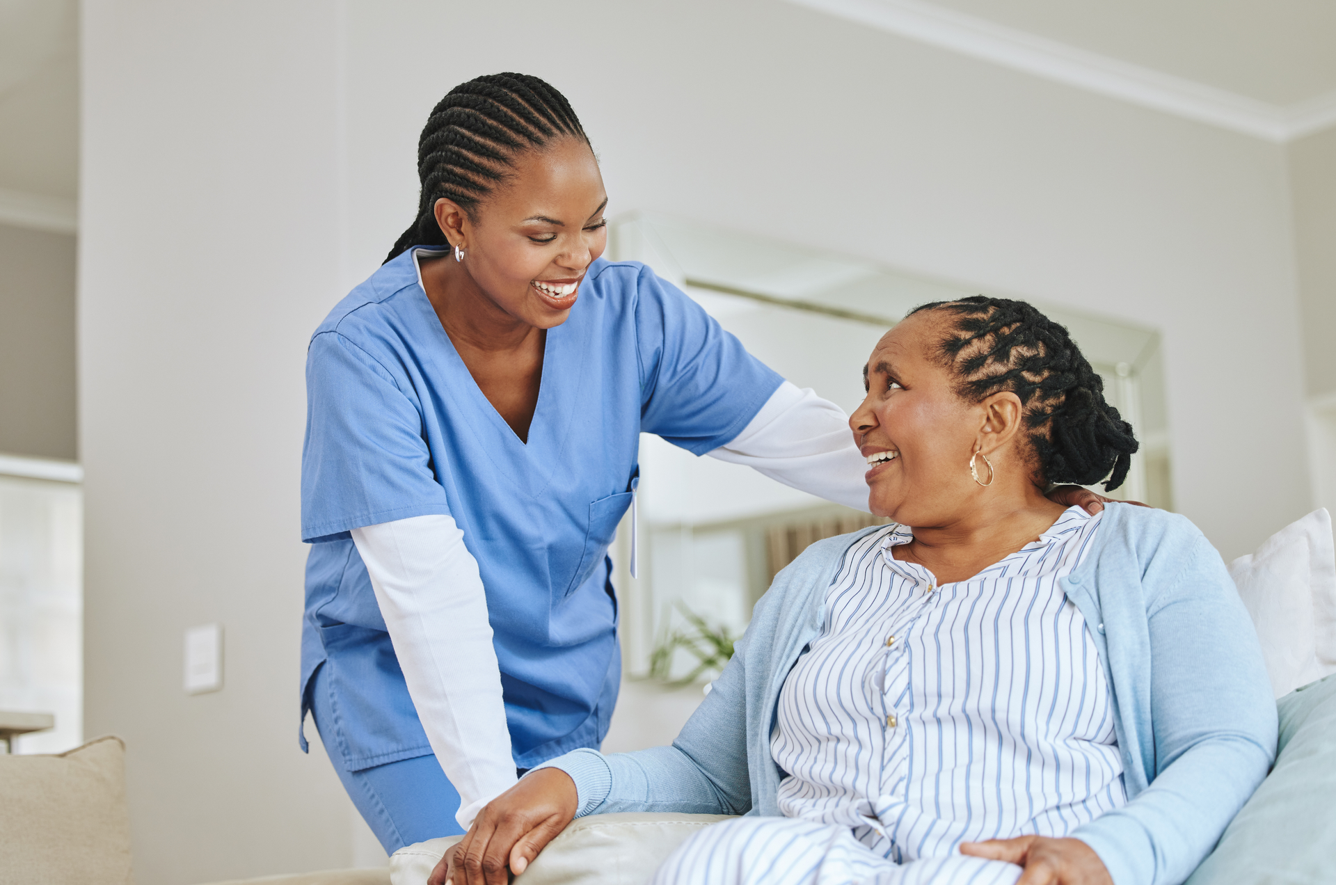 A healthcare worker in blue scrubs smiles at a woman in a light blue cardigan, indoors.