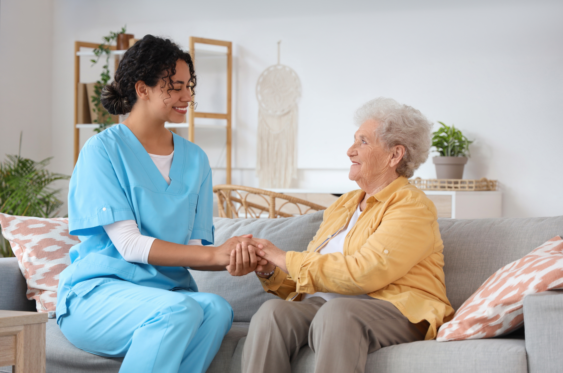 Caregiver in blue scrubs holding hands with an elderly person on a sofa, smiling.
