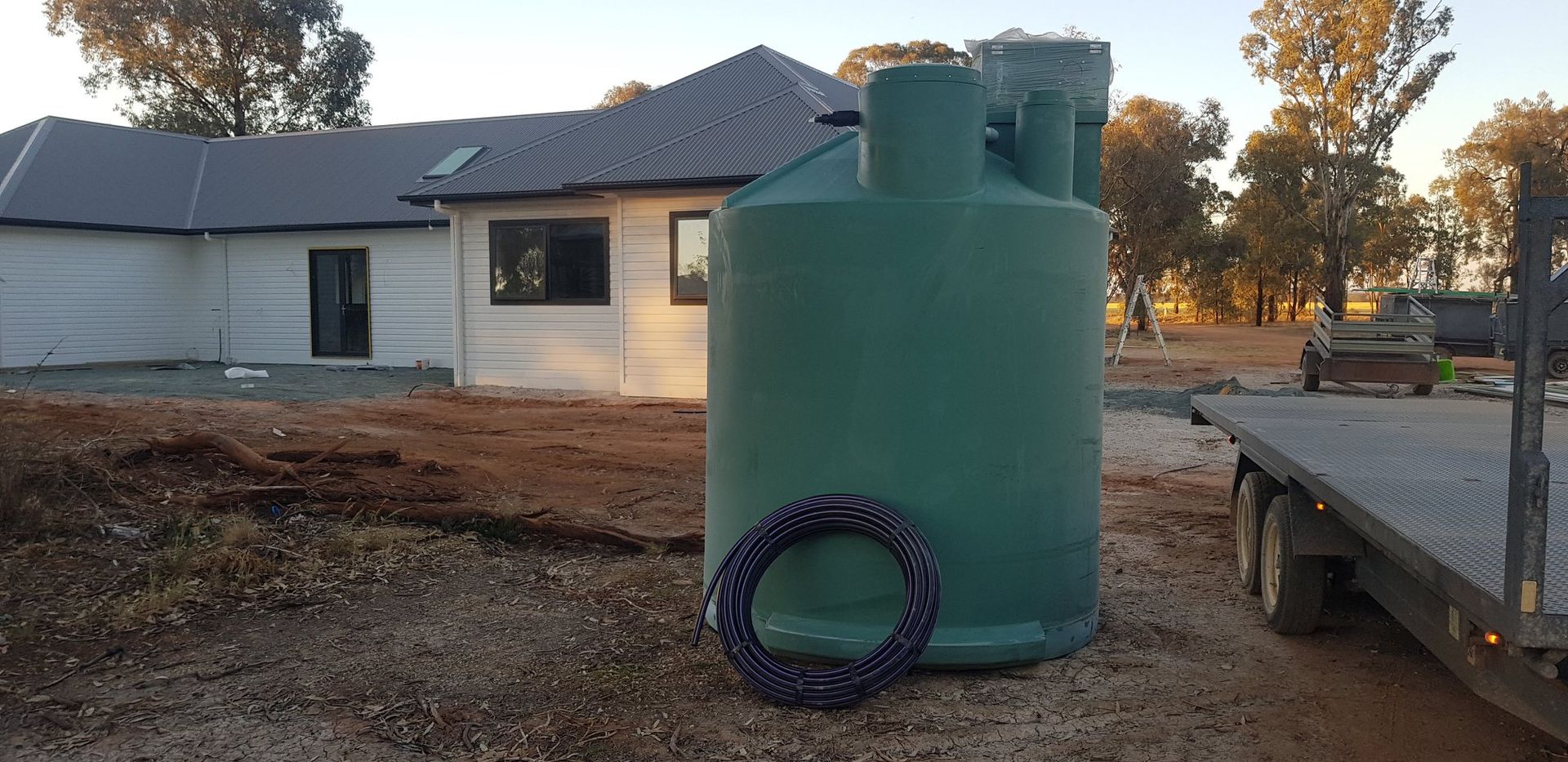 A green water tank sits near a house under construction. A trailer is nearby. Outdoors, daytime. — Mi Plumber Wagga Wagga in Temora, NSW