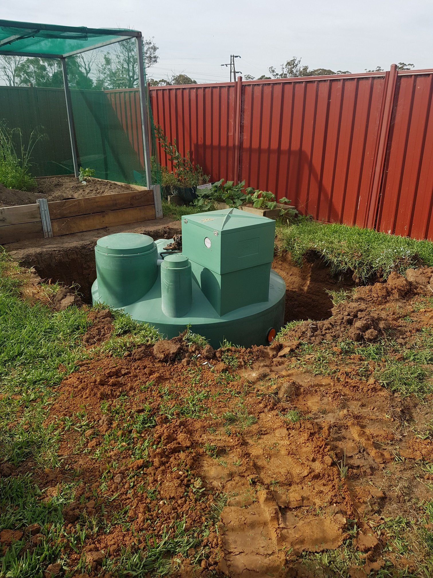 Green septic tank installed in a dirt hole, next to a red fence and garden. — Mi Plumber Wagga Wagga in Tumut, NSW