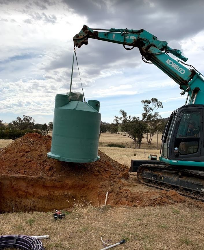 Excavator lifting a green water tank above a hole in a grassy field under a cloudy sky. — Mi Plumber Wagga Wagga in Young, NSW