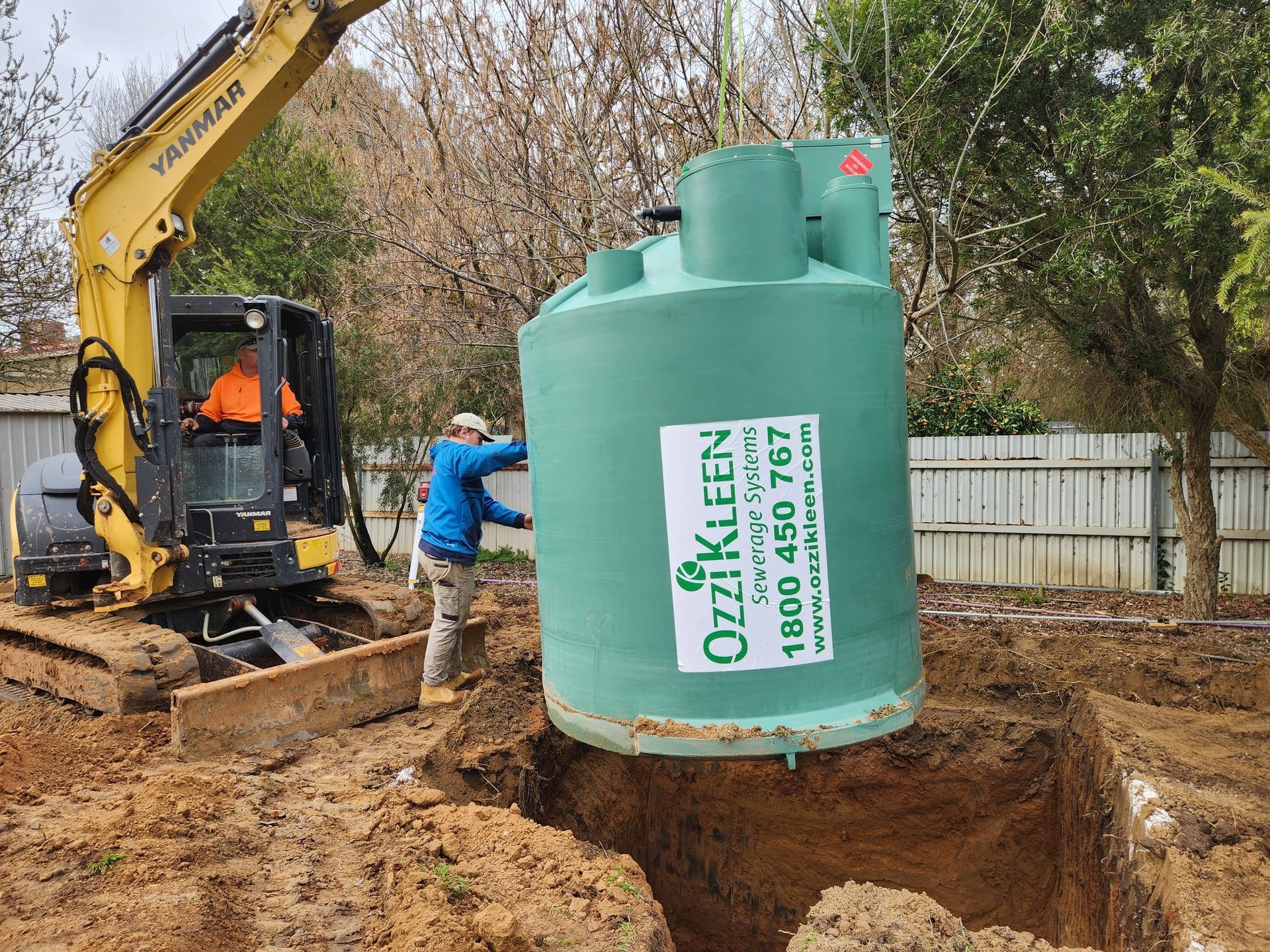 An excavator and a worker installing a green septic tank in a muddy yard. — Mi Plumber Wagga Wagga in Cootamundra, NSW