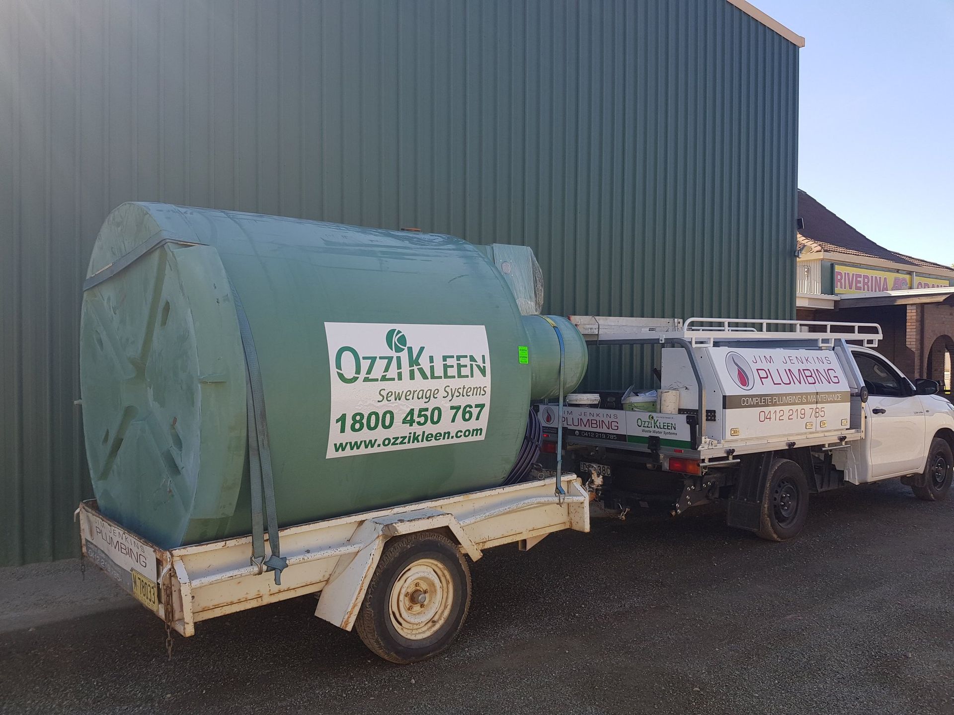 A green wastewater tank on a trailer being transported by a white truck in front of a green building. — Mi Plumber Wagga Wagga in Temora, NSW