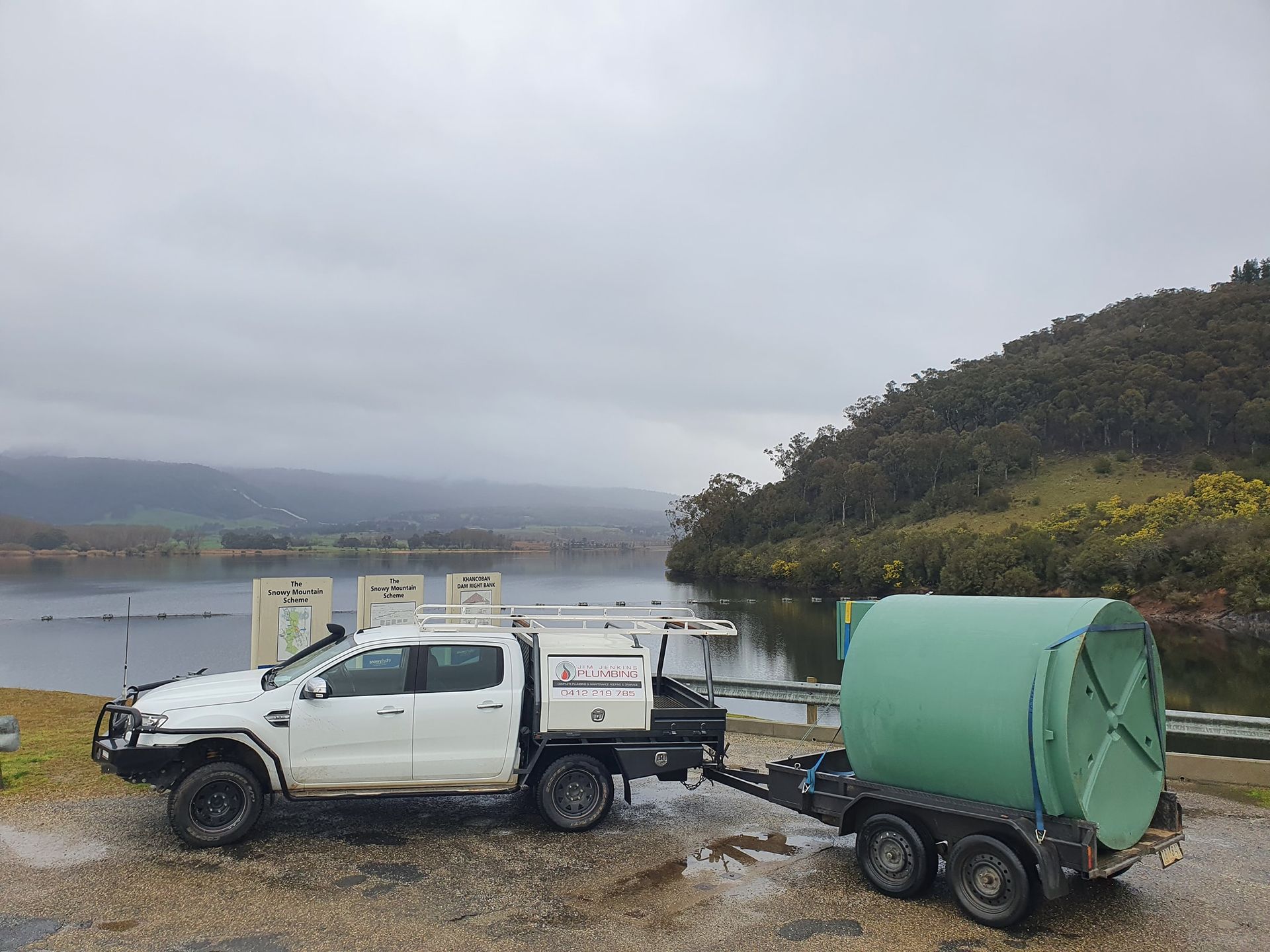White pickup truck with trailer carrying a large green cylindrical object, parked near a lake under an overcast sky. — Mi Plumber Wagga Wagga in Tumut, NSW