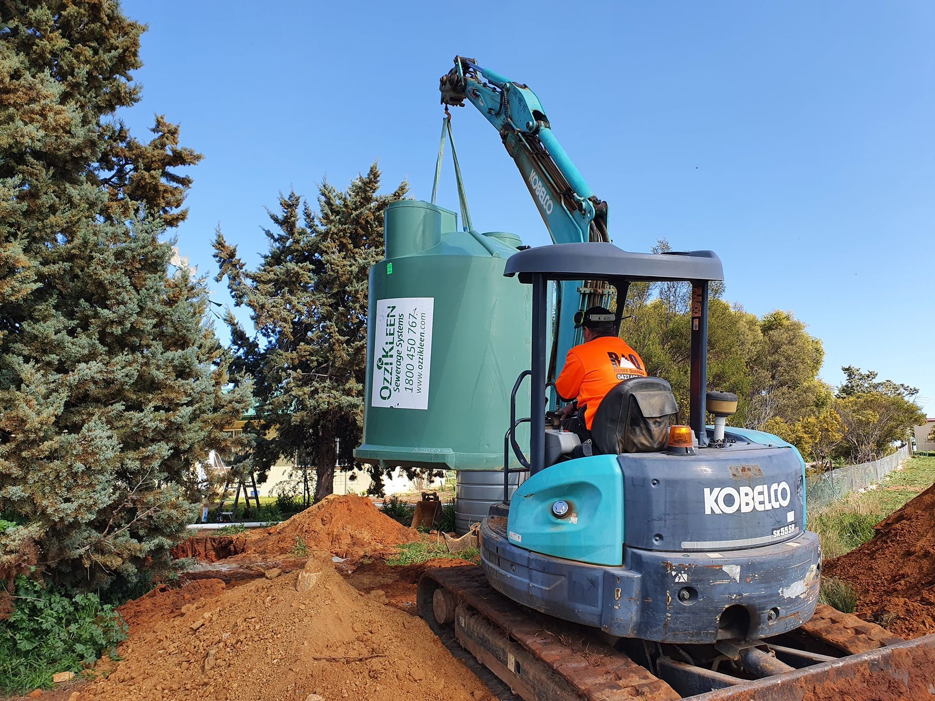 Mini excavator placing a large green tank in an excavated hole next to a tree under a blue sky. — Mi Plumber Wagga Wagga in Lake Albert, NSW
