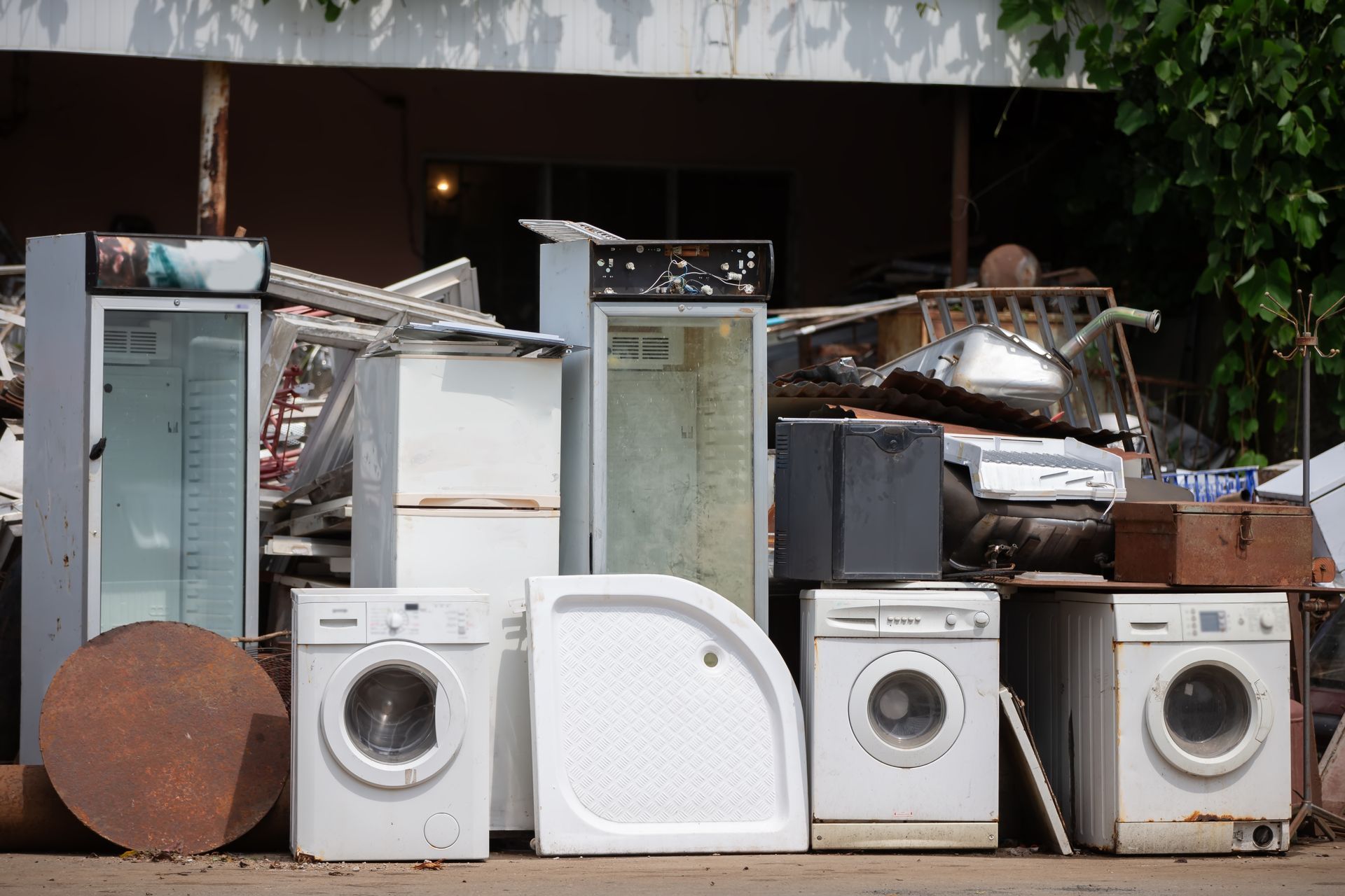 A junk pile featuring several discarded washing machines, upright glass-door fridges, and a shower base on an outdoor lot.