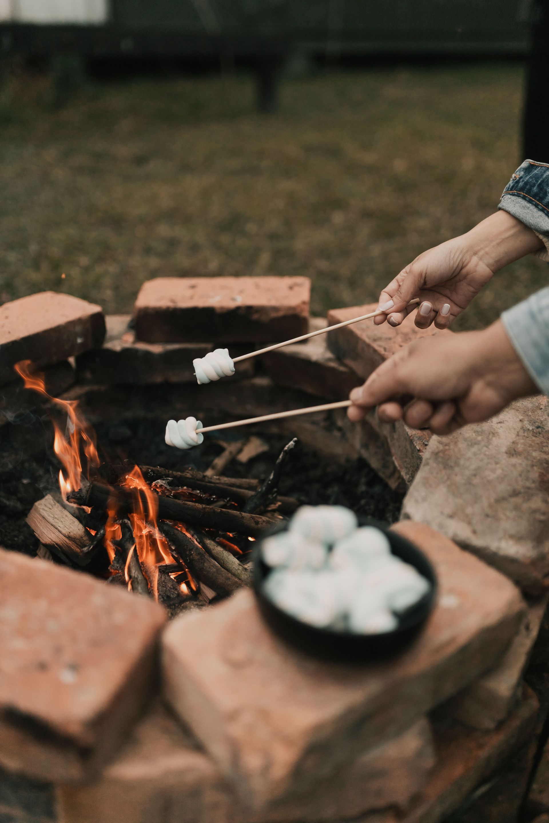 Hands roasting marshmallows over a campfire beside a brick fire pit outdoors
