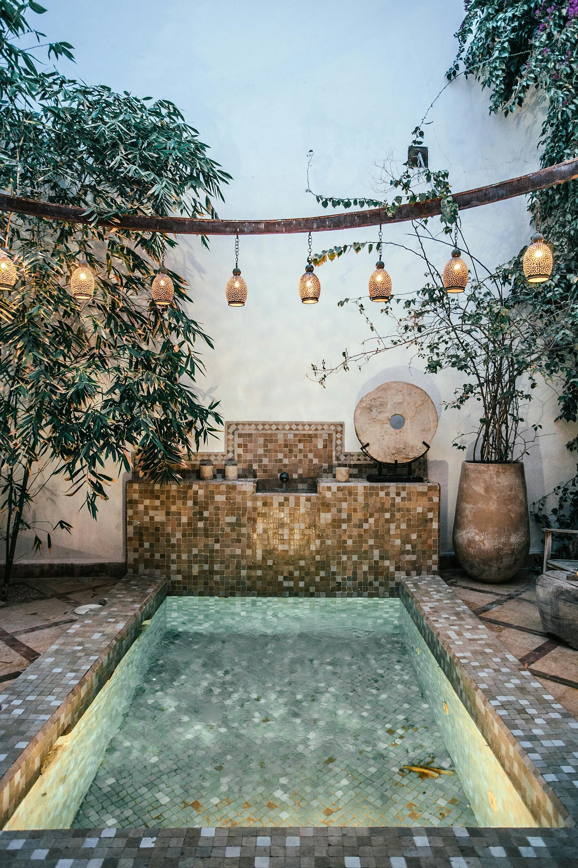 Courtyard with pool and fountain, lit with hanging lanterns and greenery, featuring stone walls and a large urn.