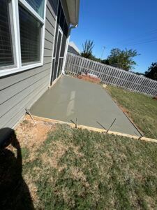 Newly poured concrete patio next to a house with gray siding and a grassy yard.