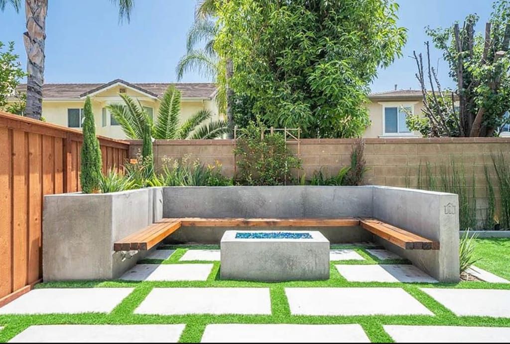 Outdoor concrete seating area with fire pit, wooden benches, and square green grass.