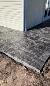 Gray stamped concrete patio next to a cream-colored building with light brown dirt in the foreground.