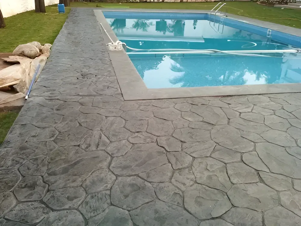 Concrete patio with stone pattern surrounds a swimming pool filled with water.