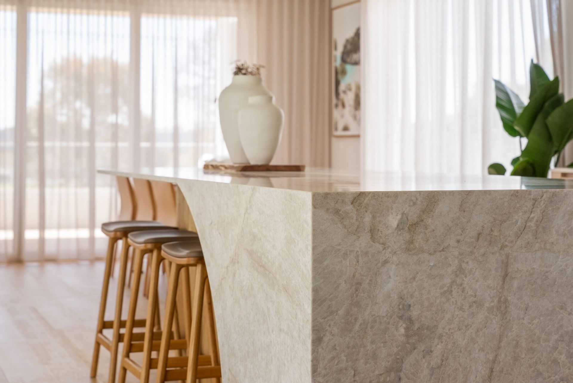 Modern kitchen island with wooden stools, a white vase, and sunlit sheer curtains