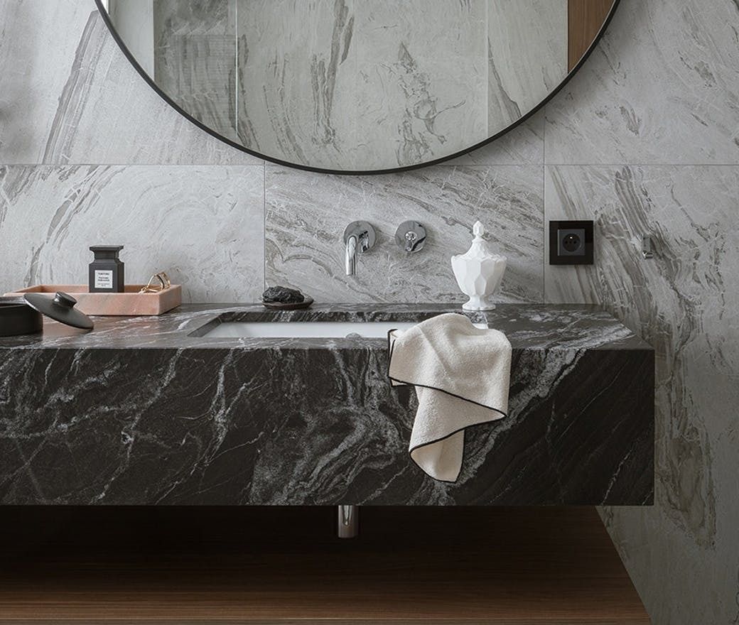 Modern marble bathroom vanity with dark sink, round mirror, and towels hanging from the counter