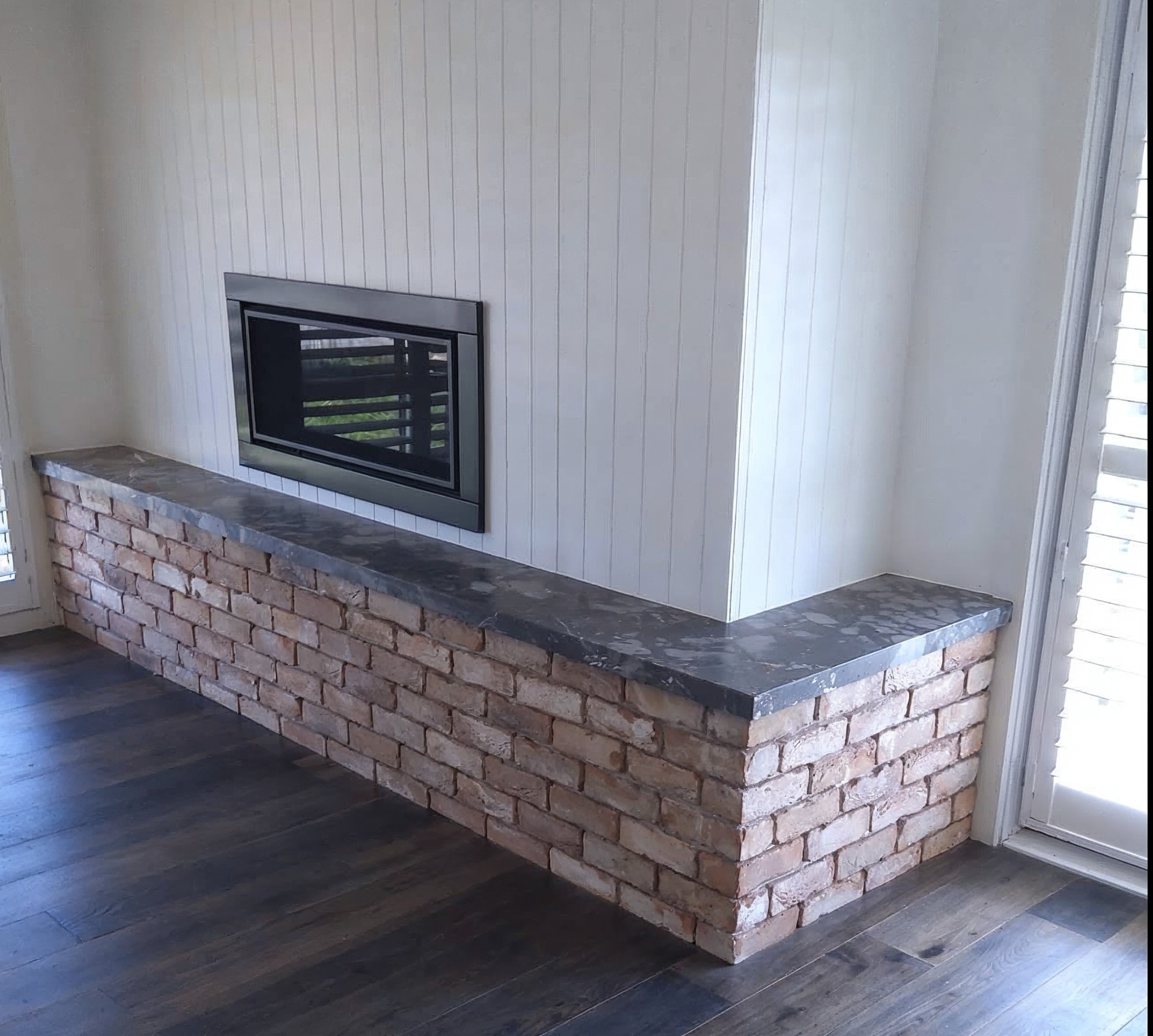 Modern living room fireplace with white walls, brick base, dark wood floor, and a large window.