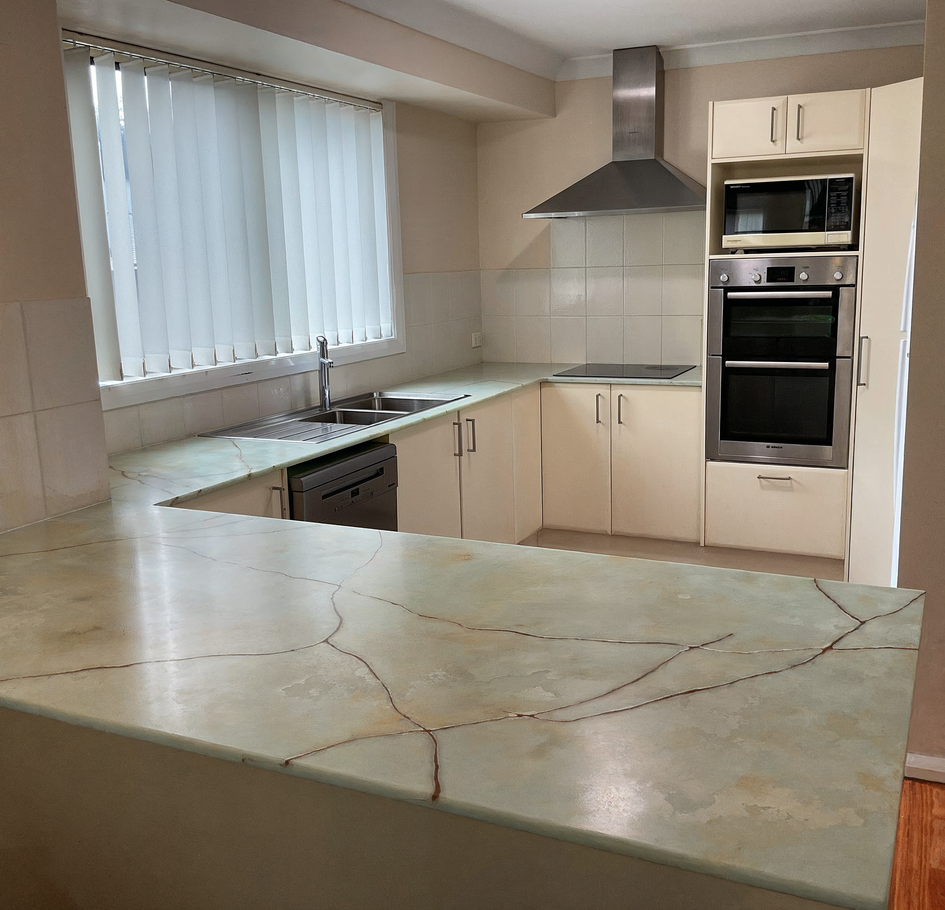 Bright modern kitchen with cream cabinets, built-in oven, stainless hood, sink by window, and marble-look island countertop