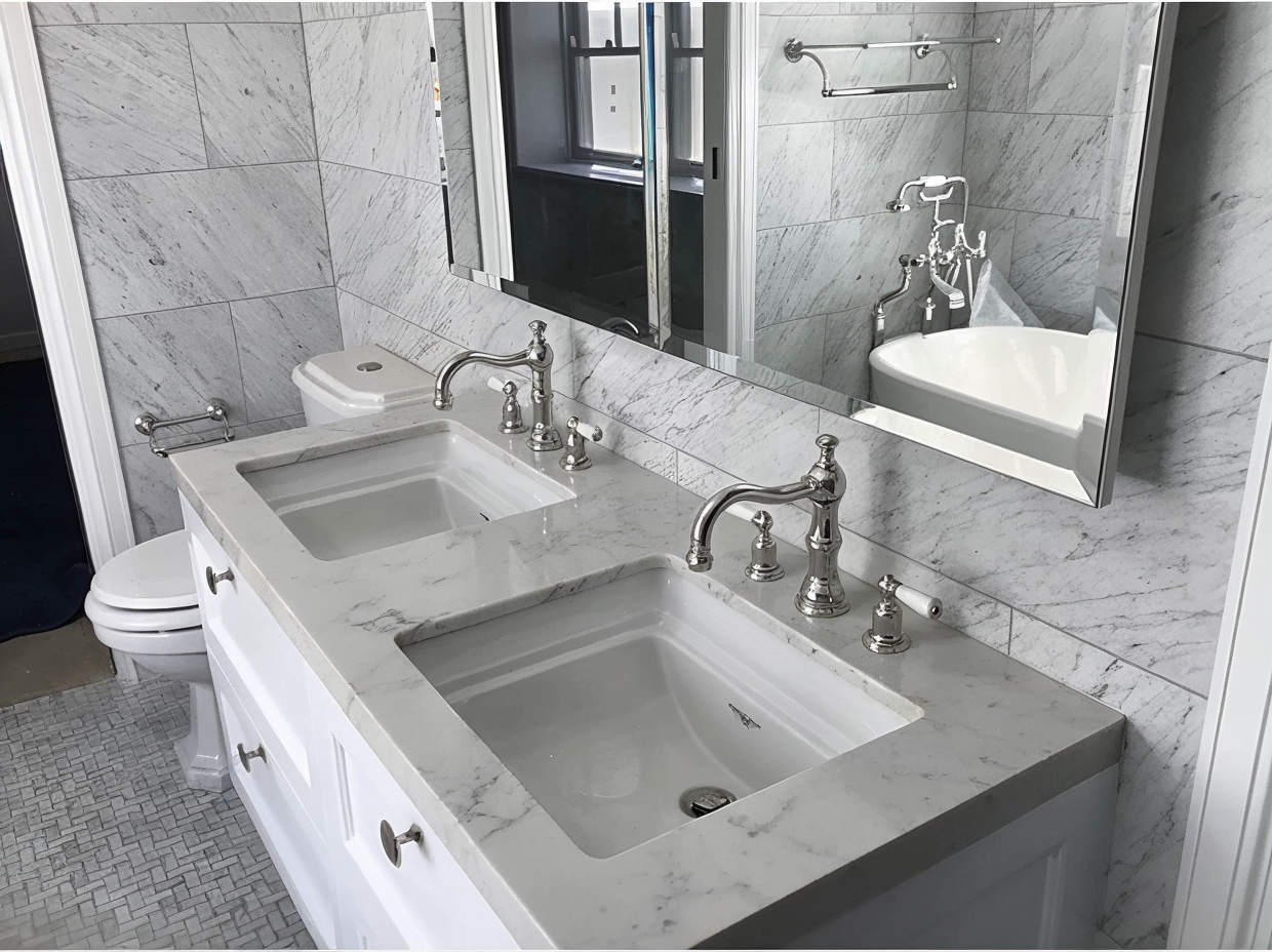White marble bathroom with double sinks, chrome faucets, mirror, and a toilet in the background