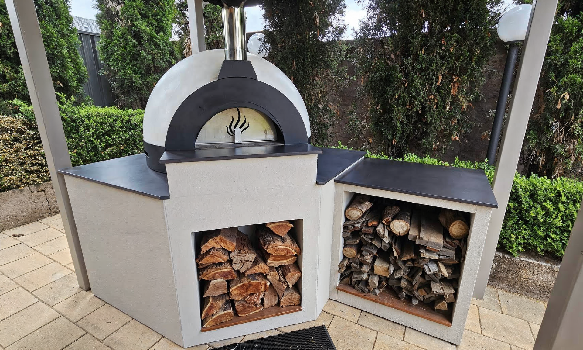 Outdoor wood-fired oven with open firewood storage under a black-and-white canopy in a garden setting