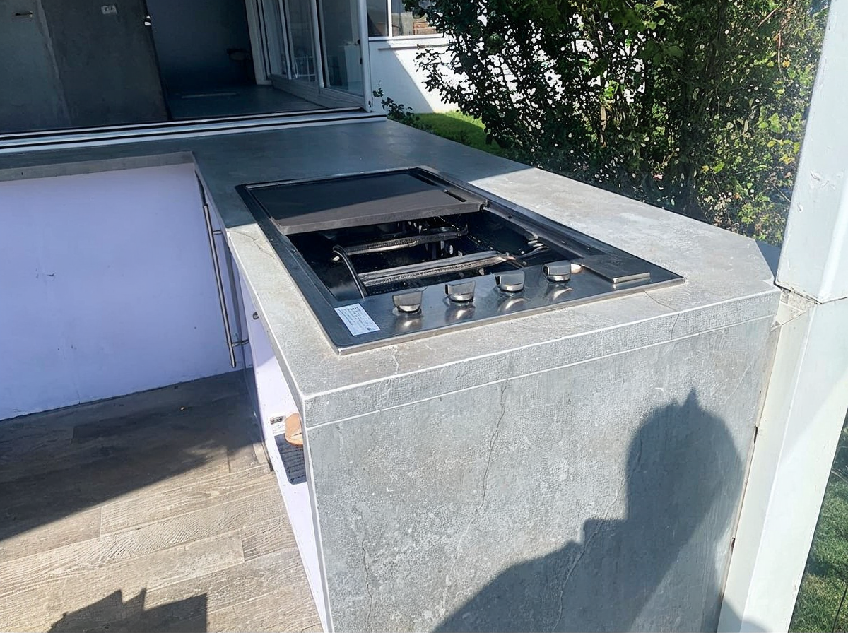 Outdoor stainless-steel gas grill on a patio beside a building and fence, with a shadow on the side.