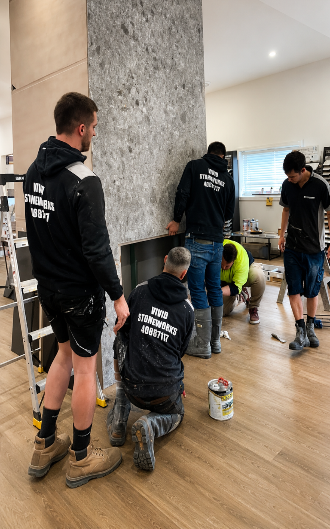Workers painting a wall in a bright room with wood floors, one kneeling near paint cans.