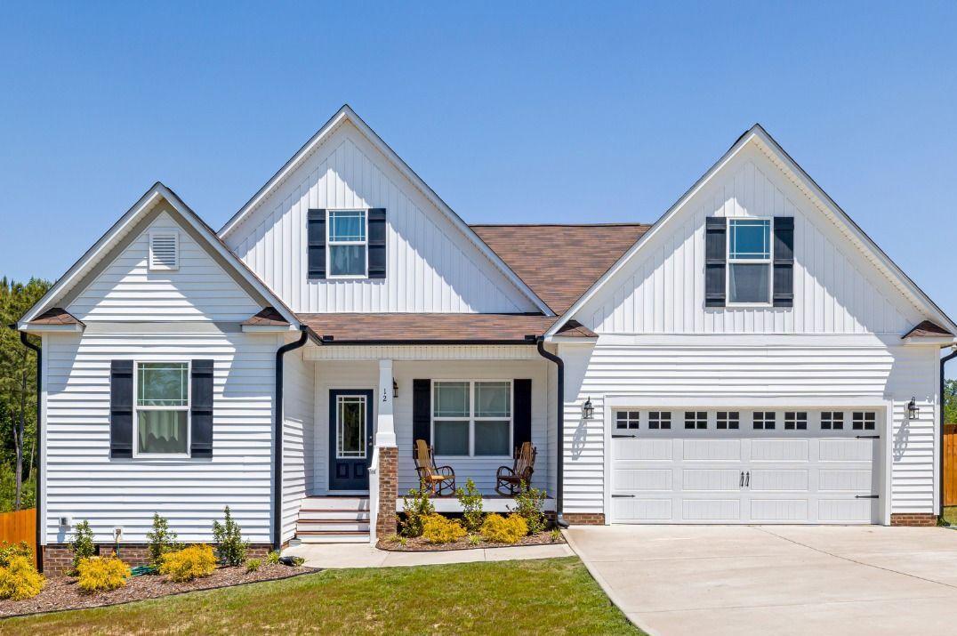 Single-story white house with black shutters, brown roof, double garage, and a small front porch with chairs and landscaping