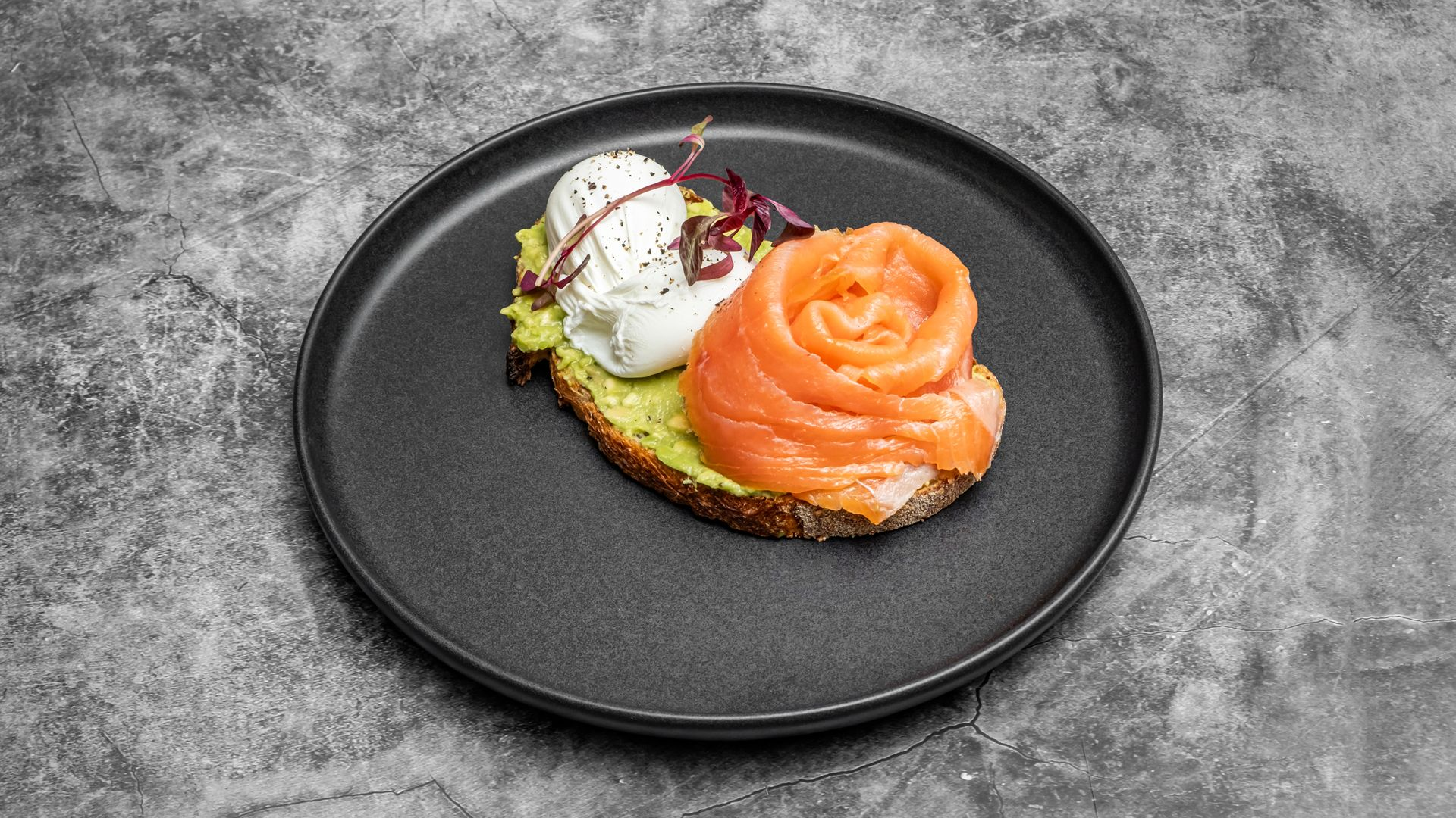 Avocado toast topped with a poached egg and a salmon rose, on a black plate.