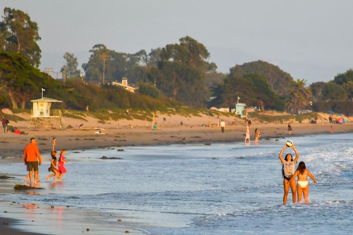 People playing on a sandy beach; several are in the water. A lifeguard station and trees are in the background.