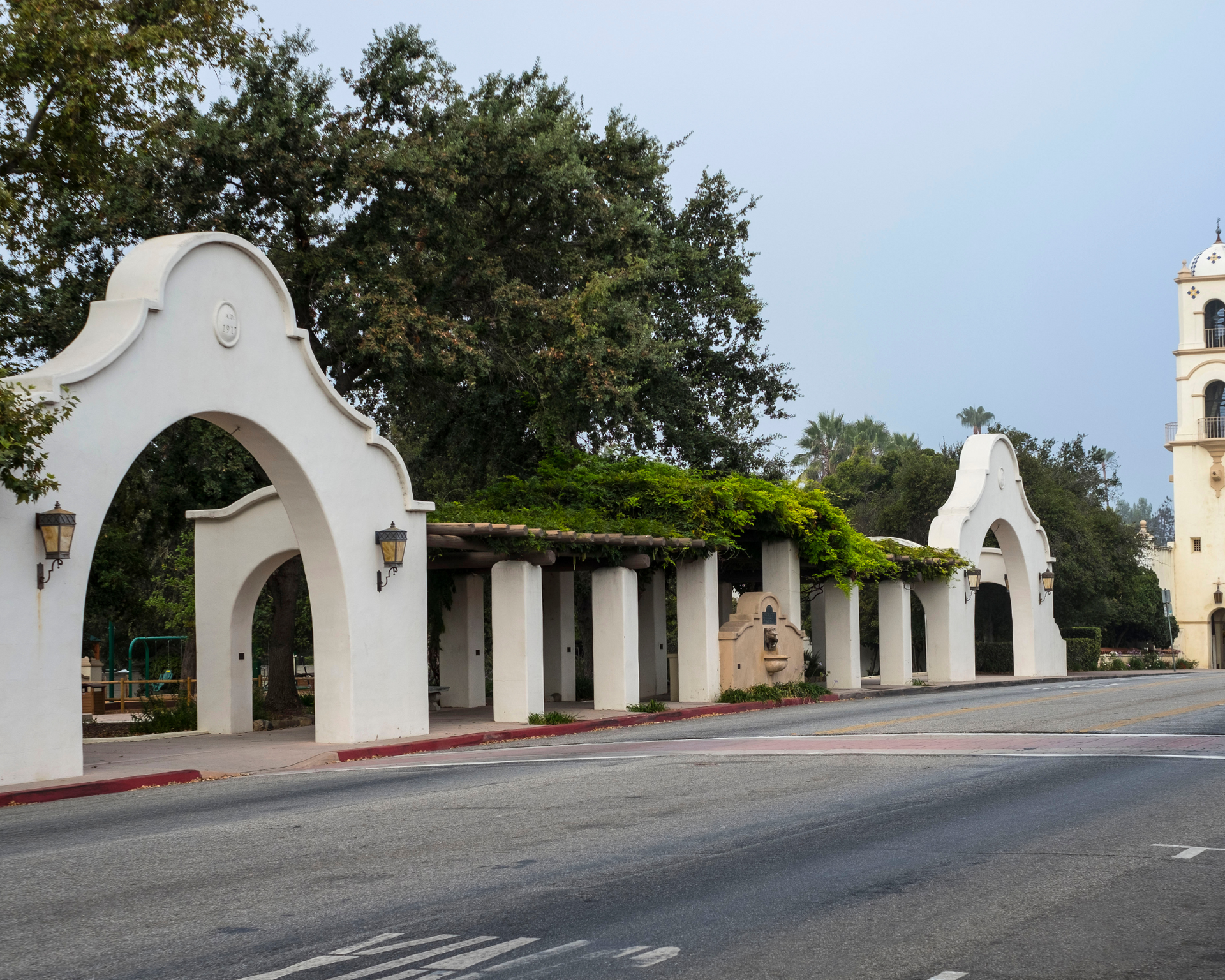 White arched entrance with green vines, adjacent to a tower, along a street.