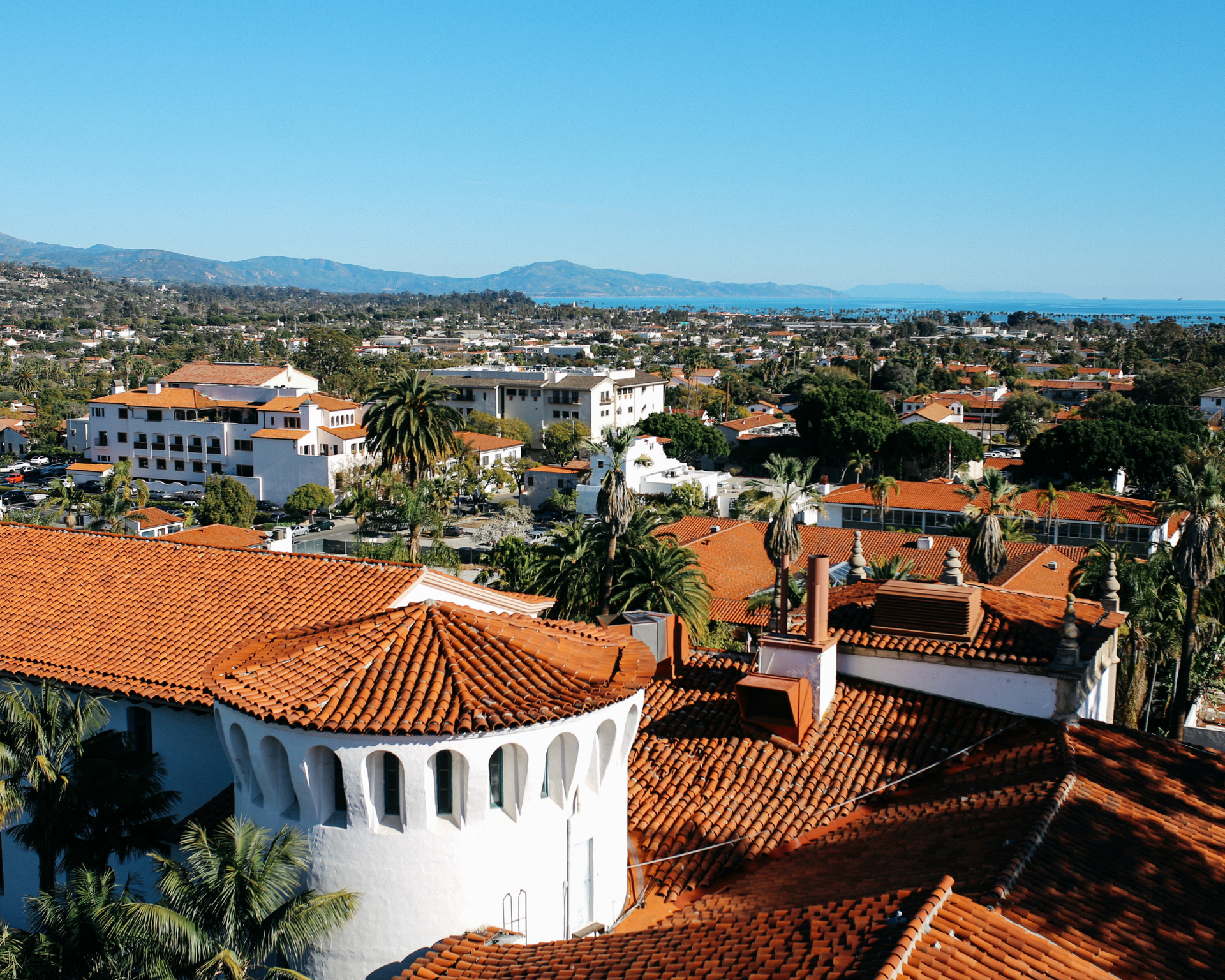 View of Santa Barbara, California, with red-tile roofs, white buildings, palm trees, mountains, and the ocean under a clear blue sky.