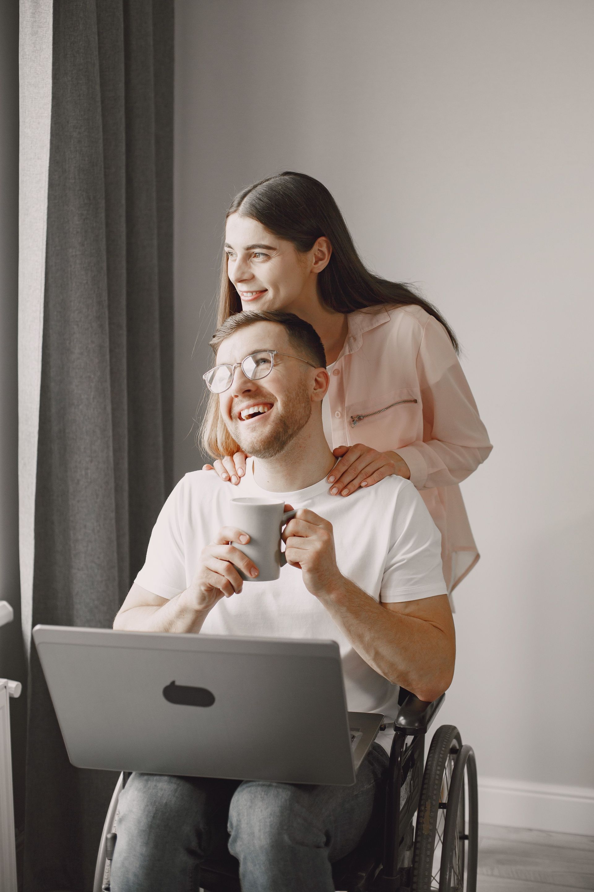 A young man using a wheelchair laughs as a young woman rests her hands on his shoulders