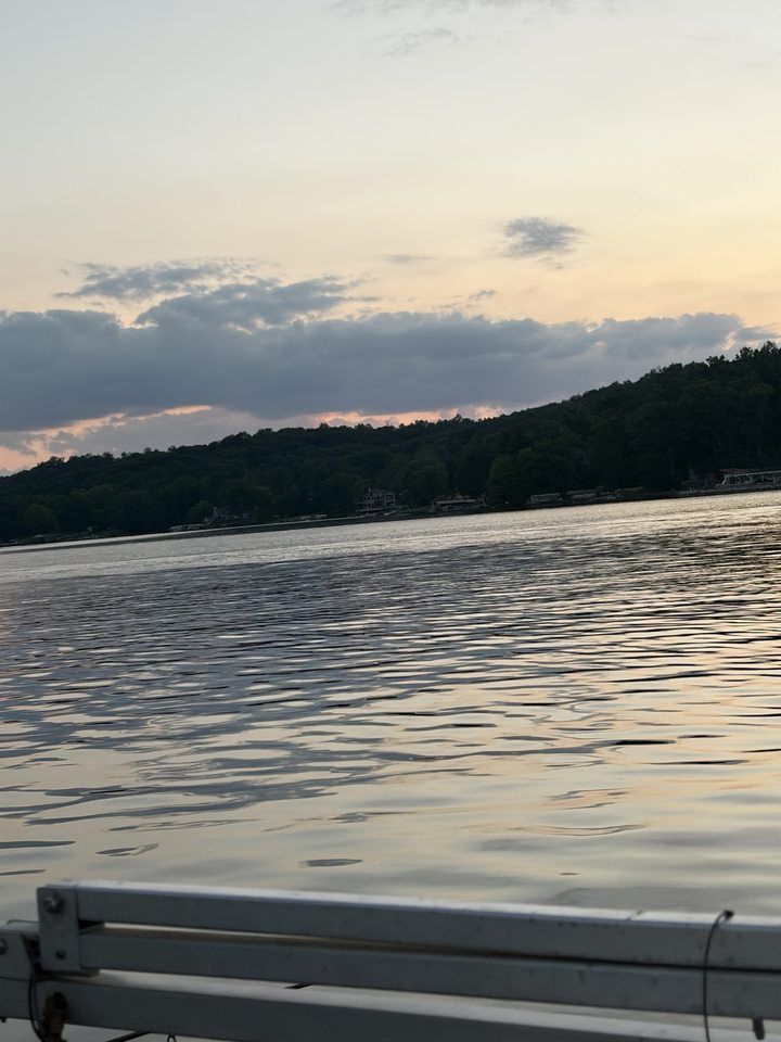 Calm lake at sunset with tree-lined shore and a cloudy sky, seen over a boat railing