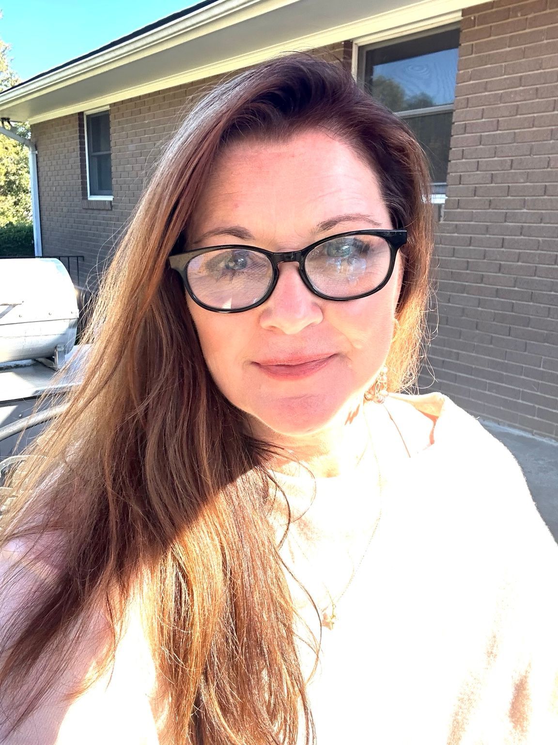 Woman with glasses outdoors in bright sunlight, standing near a brick house.