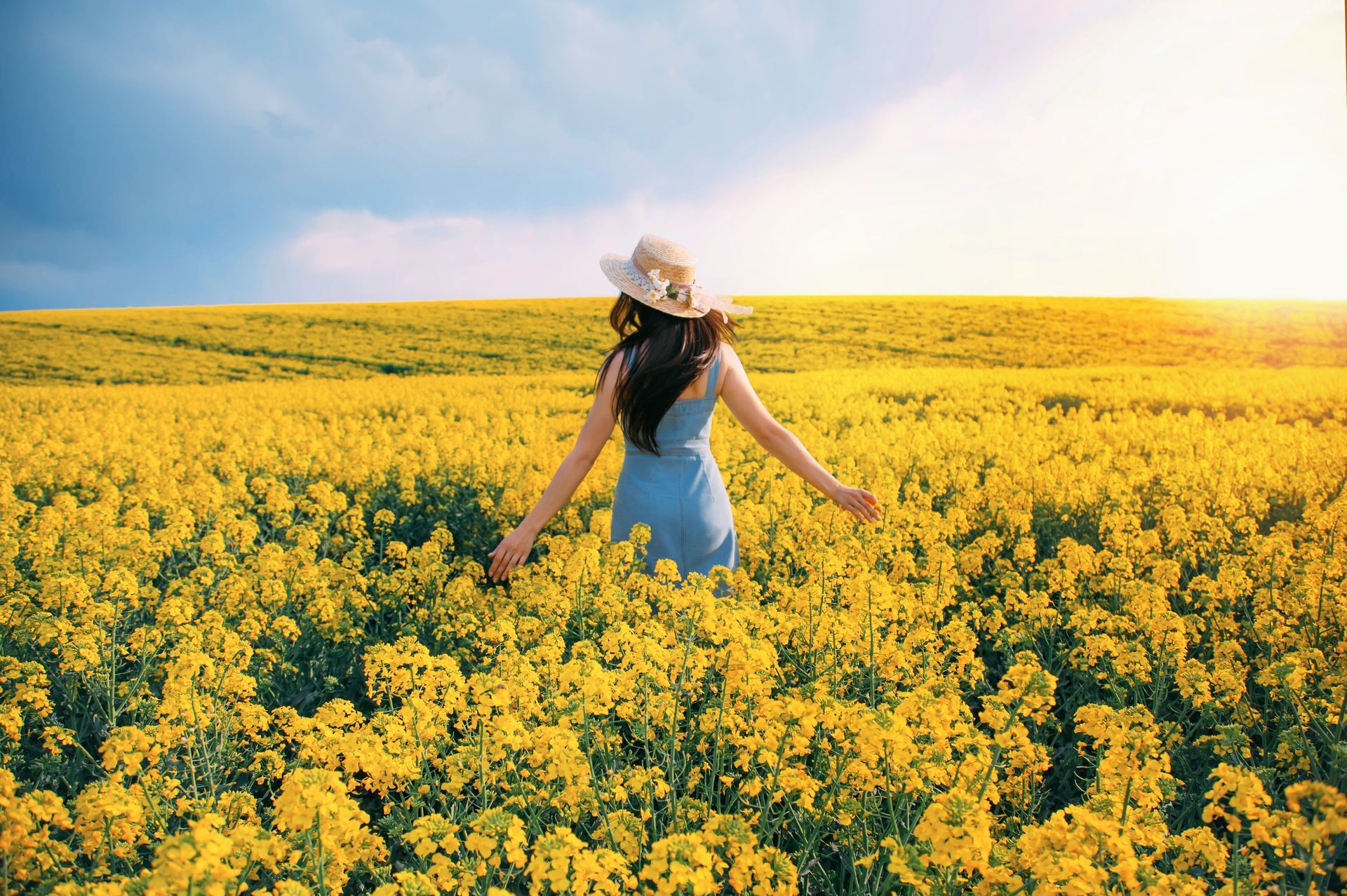 Woman in a white dress and hat standing in a bright yellow flower field under a blue sky