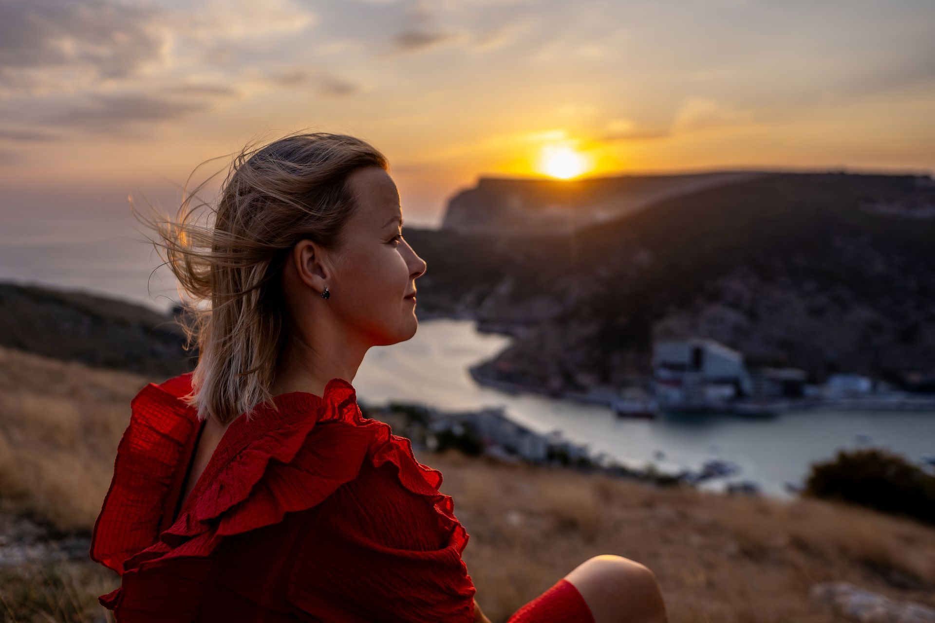 Woman in a red dress overlooking a sunset over a coastal bay and hillside village