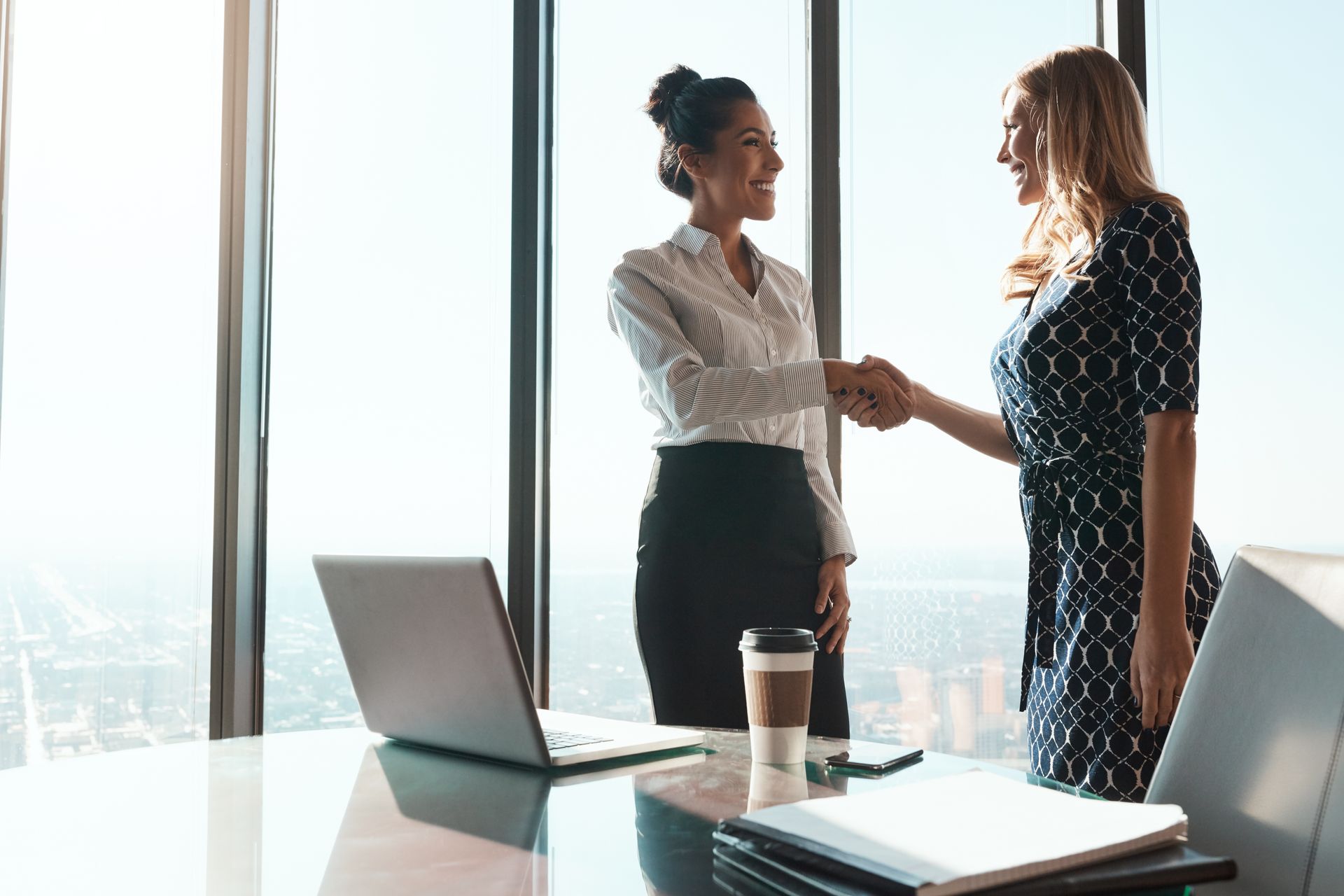 Two coworkers shake hands in a bright office beside a laptop and coffee cup.