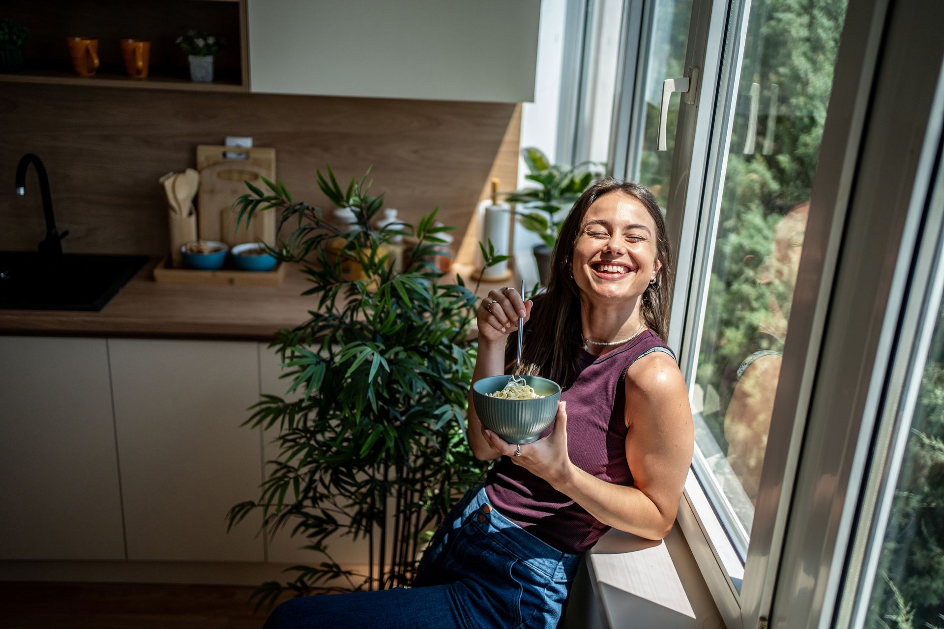 Woman smiling and holding a bowl in a sunlit kitchen by a window.